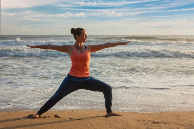 mulher-fazendo-yoga-asana-virabhadrasana-1-pose-de-guerreiro-na-praia_163782-1479